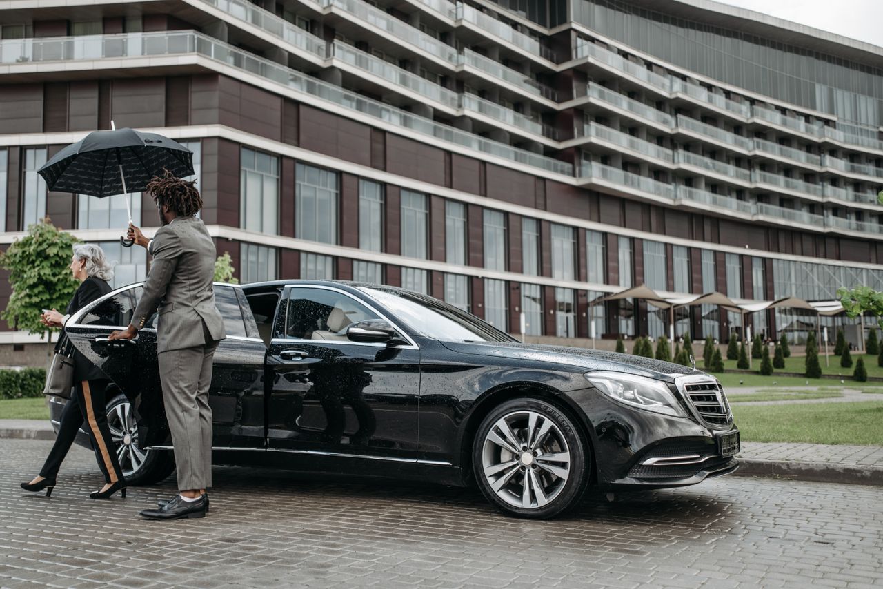 A business professional exits a luxury car while being shielded by an umbrella.