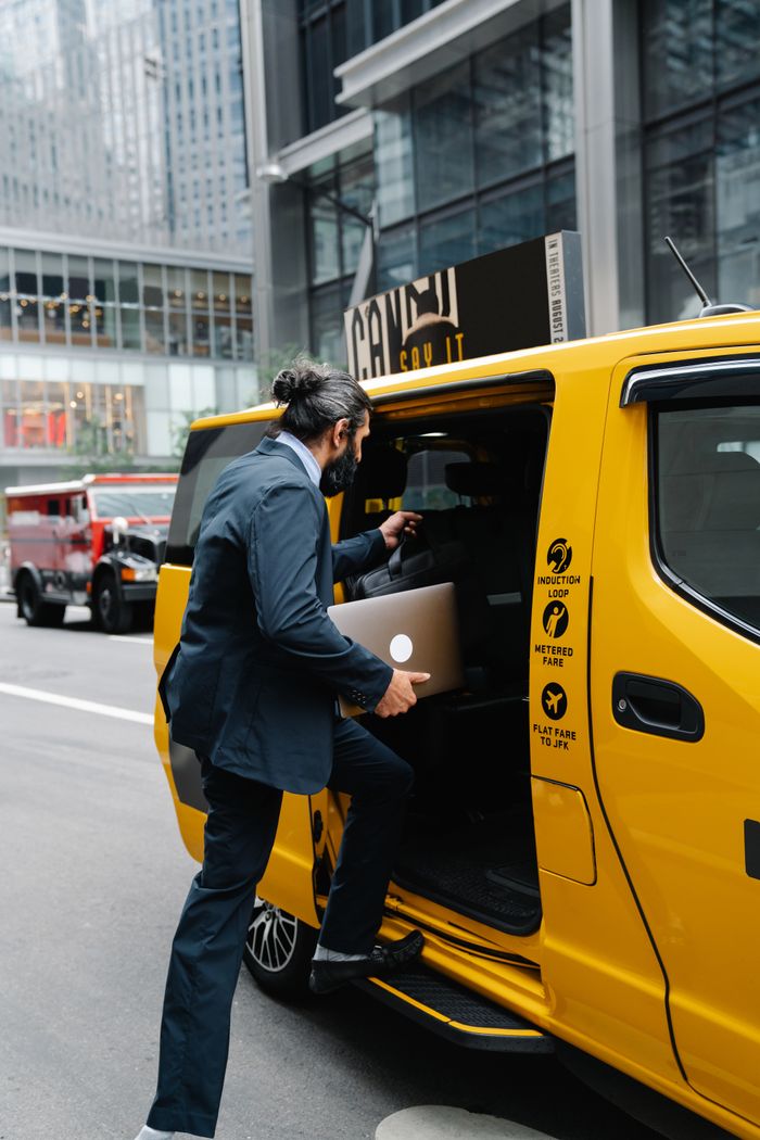A businessman in formal attire enters a yellow taxi in a bustling city area, holding a laptop.